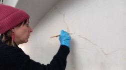 Eine Frau hat einen Pinsel in der rechten Hand. Mit diesem f&auml;hrt sie &uuml;ber einen dunklen Strich auf einer wei&szlig;en Wandfl&auml;che.