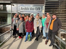 Foto: 16 Frauen und Männer stehen vor einer Tür auf der steht „Archiv des LVR“ und lächeln in die Kamera