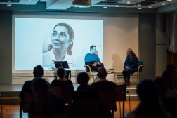 Foto: Auf dem Foto sind ein Mann und eine Frau zu sehen, die vor Publikum auf einer Bühne sitzen und sich unterhalten. Auf einer Leinwand das Foto einer Frau.