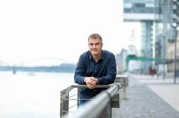 Ein Mann mit Brille und blauem Hemd lehnt sich auf ein Geländer und blickt in die Kamera. Im Hintergrund sind verschwommen das Rheinufer und die Rheinbrücke zu sehen.